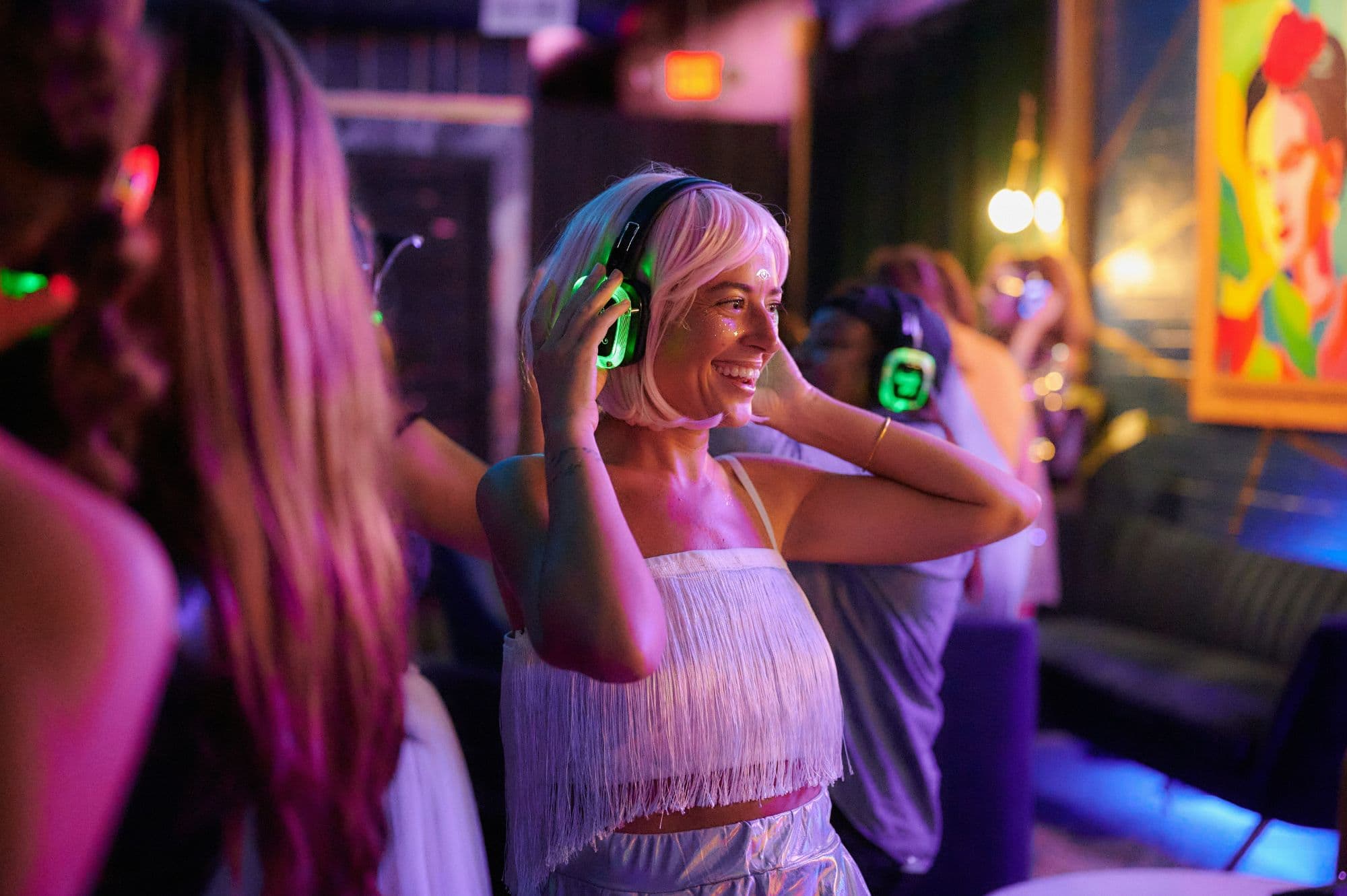 A guest at a silent disco smiling as she adjusts her green-lit wireless headphones, with others dancing behind her